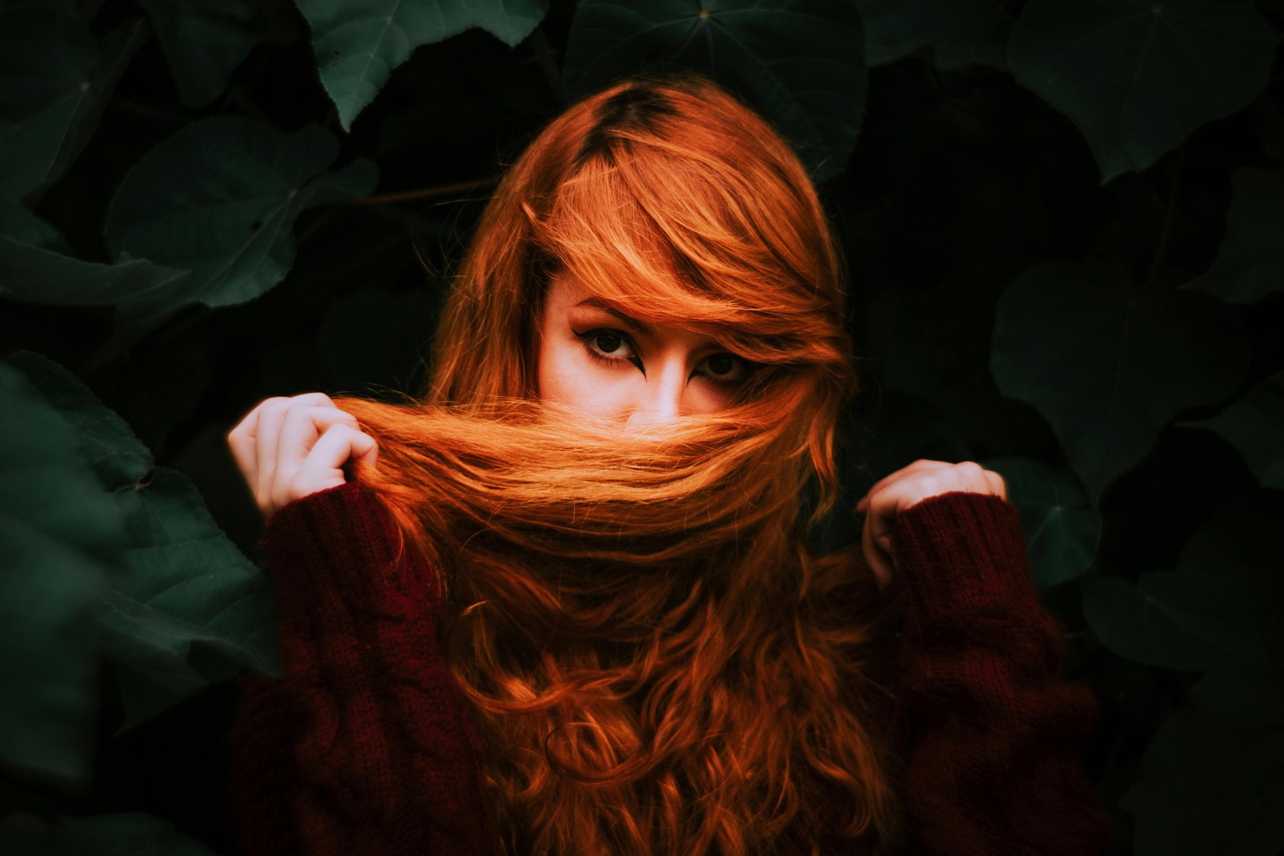 A captivating portrait of a woman with auburn hair partially covering her face, surrounded by lush green leaves. Portrait captivant d'une femme aux cheveux auburn couvrant partiellement son visage, entourée de feuilles vertes luxuriantes.