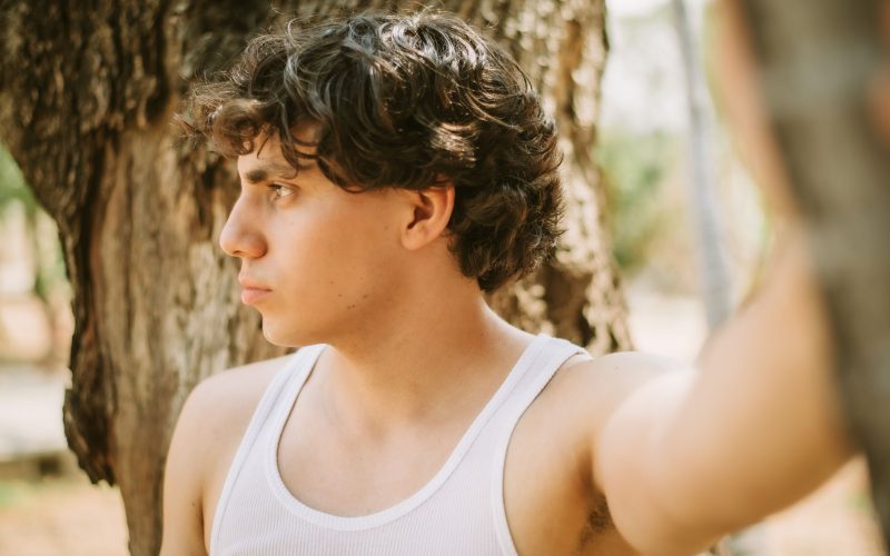 Jeune homme aux cheveux ondulés vu de profil, en débardeur sous un arbre