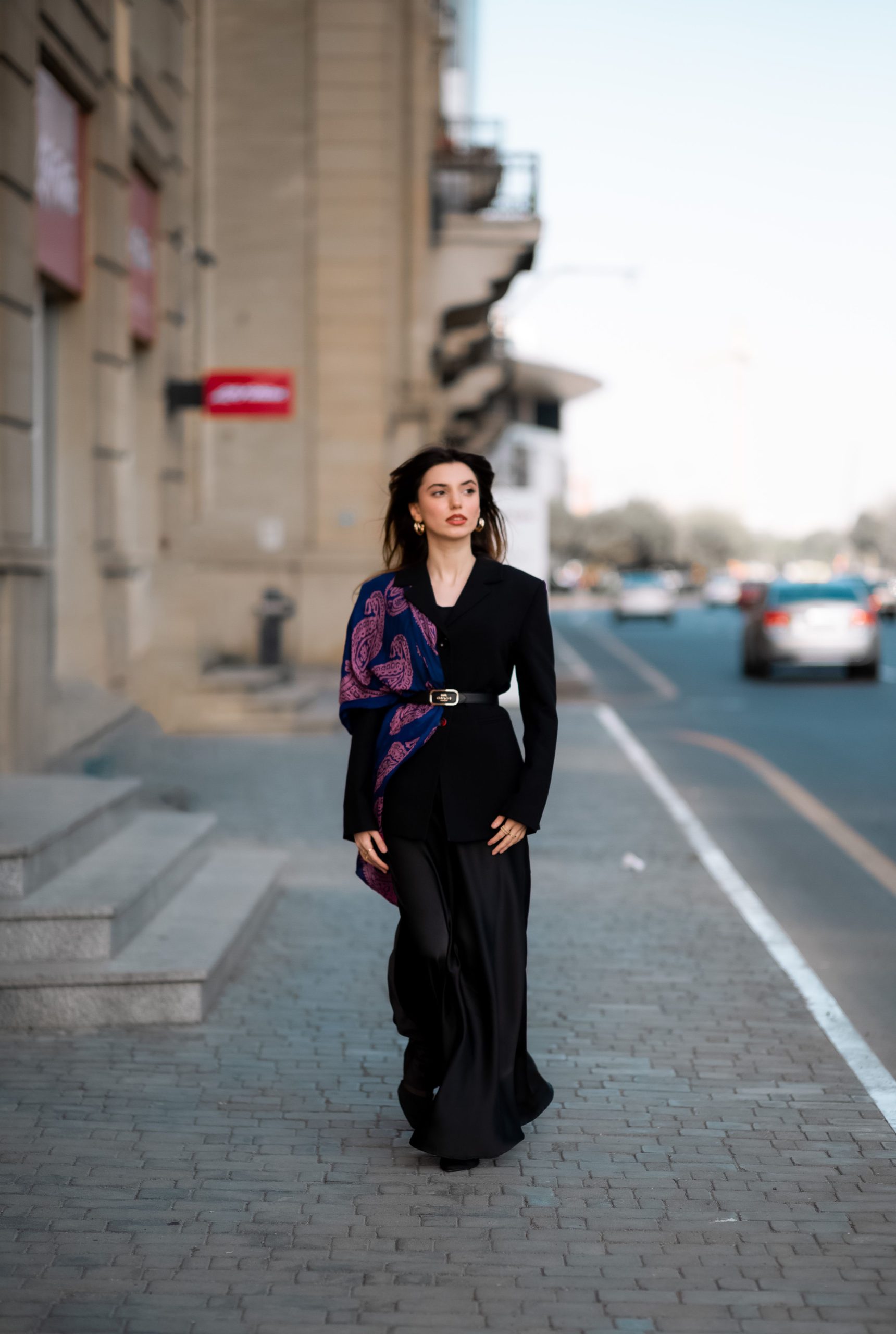 Elegant woman in black attire walking confidently in a city street setting, showcasing urban fashion style. Femme élégante en tenue noire marchant avec assurance dans une rue de la ville, mettant en avant un style de mode urbaine.