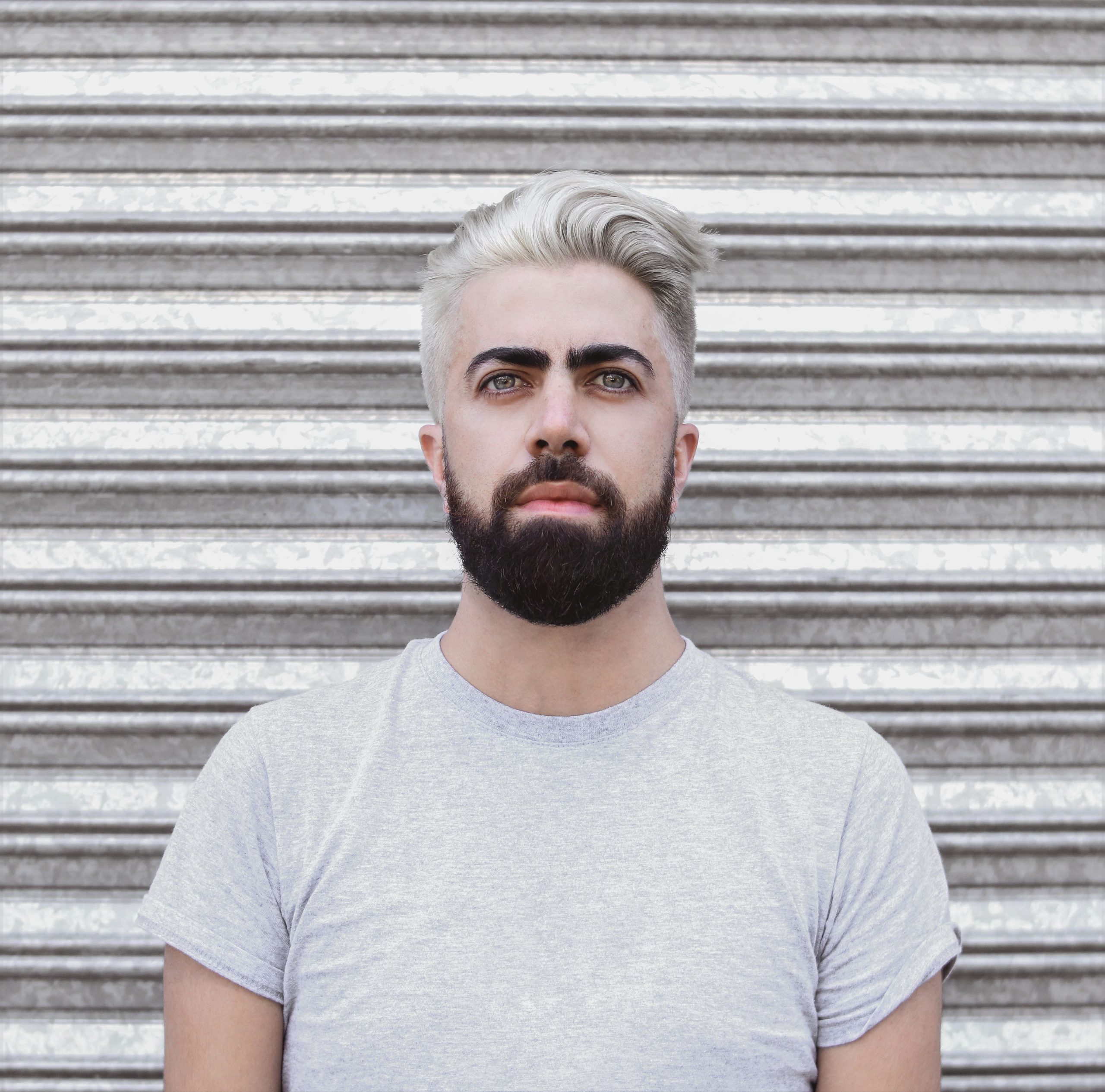 Close-up portrait of a man with a beard and gray hair against a striped metal background. Portrait en gros plan d'un homme barbu aux cheveux gris sur un fond métallique rayé.