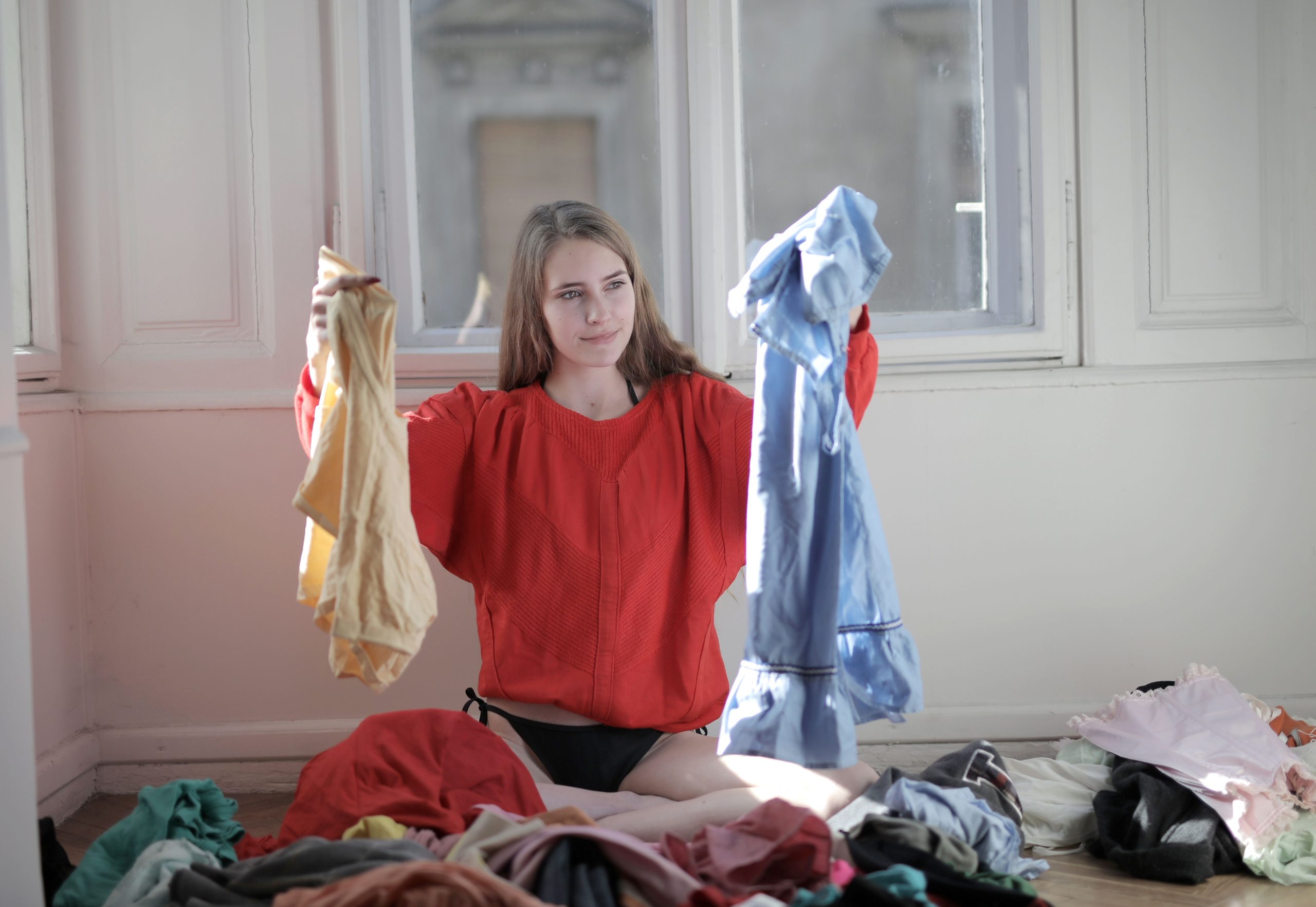 A woman in a red sweater deciding between clothes in a messy room. Une femme en pull rouge décidant entre des vêtements dans une chambre en désordre.