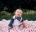 A happy baby sitting on a cherry-printed blanket outdoors, enjoying a sunny day. Un bébé heureux assis sur une couverture imprimée de cerises à l'extérieur, profitant d'une journée ensoleillée.