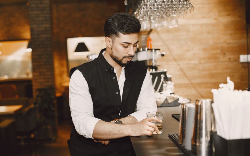 Homme en chemise blanche et gilet noir accoudé à un bar le soir