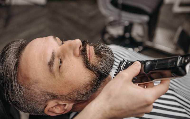 Barbe courte poivre et sel taillée à la tondeuse électrique, homme allongé dans un fauteuil de barbier