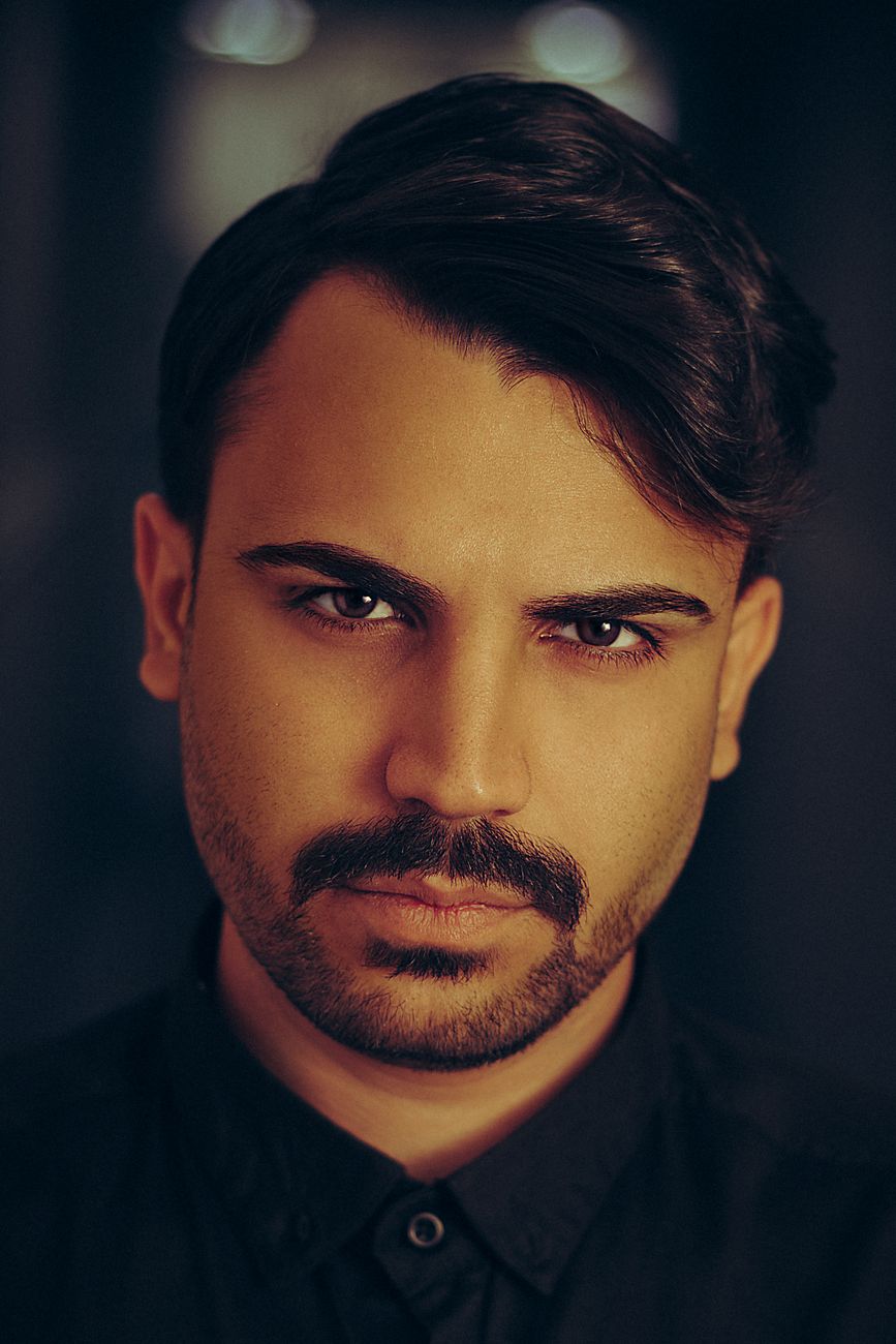 Homme à la moustache dessinée et à la barbe courte, de face sur fond sombre