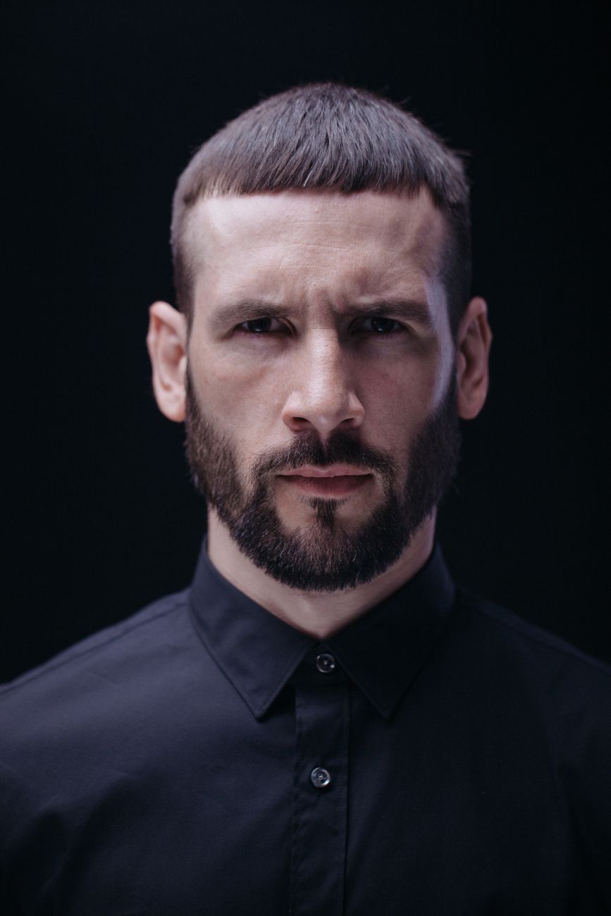 Homme à la barbe courte brun foncé, uniforme, de face sur fond noir