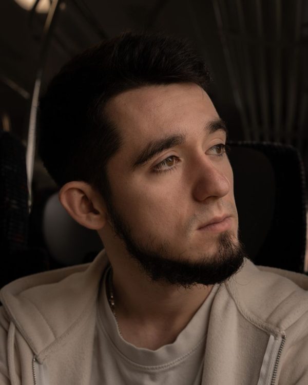 Jeune homme de trois quarts, collier de barbe brun qui suit la mâchoire, joues rasées