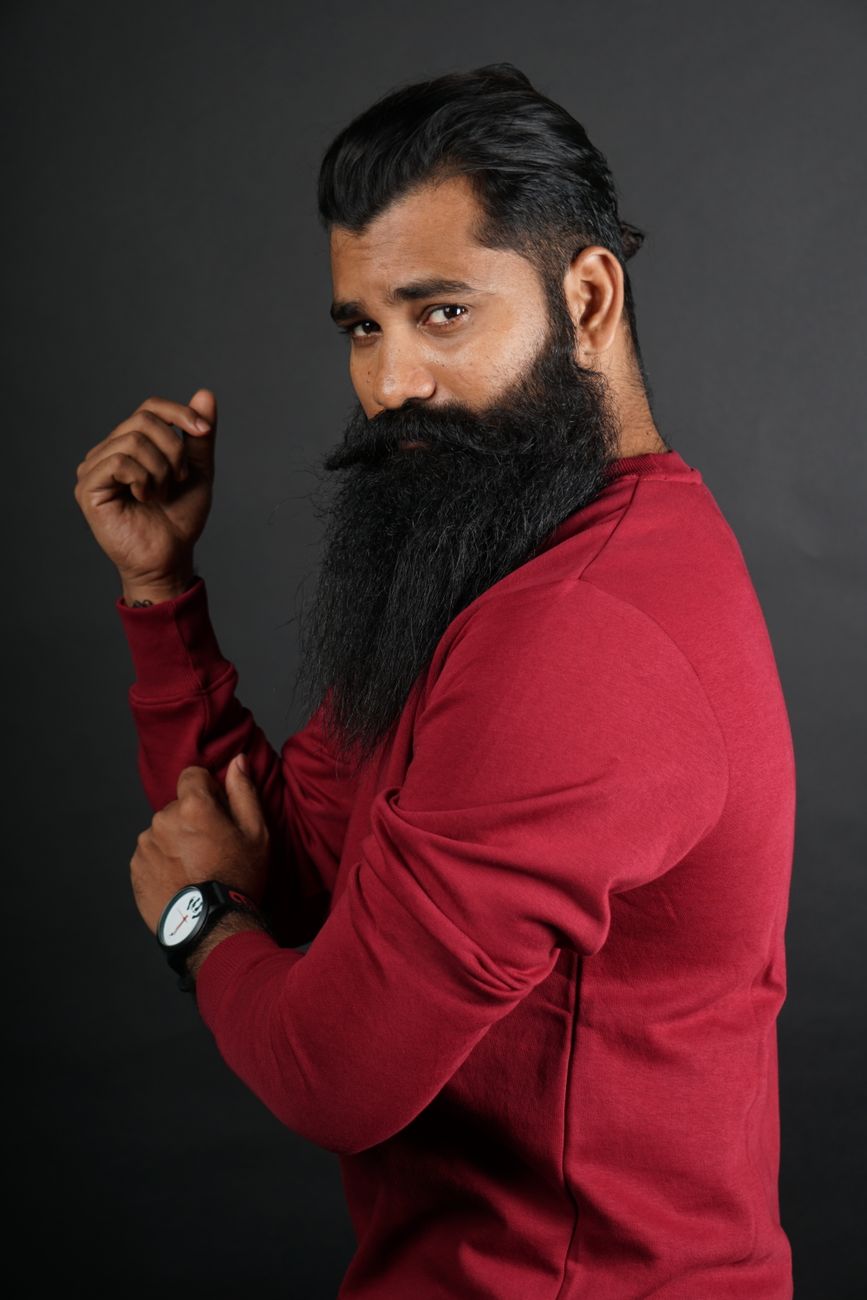 Homme à la barbe noire longue et fournie, pull rouge, sur fond sombre