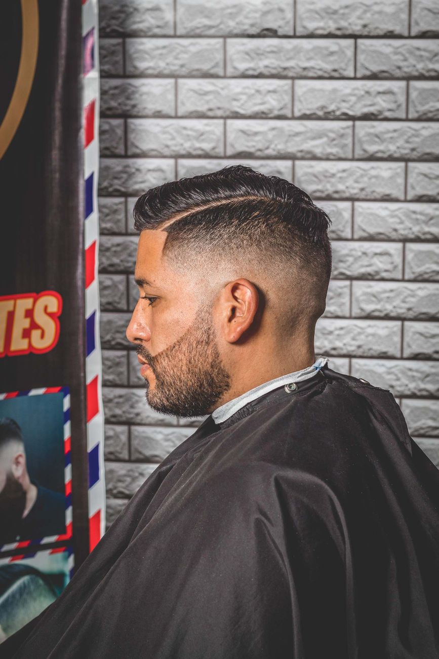 Homme barbu de profil, raie marquée et côtés en dégradé, cape de barbier